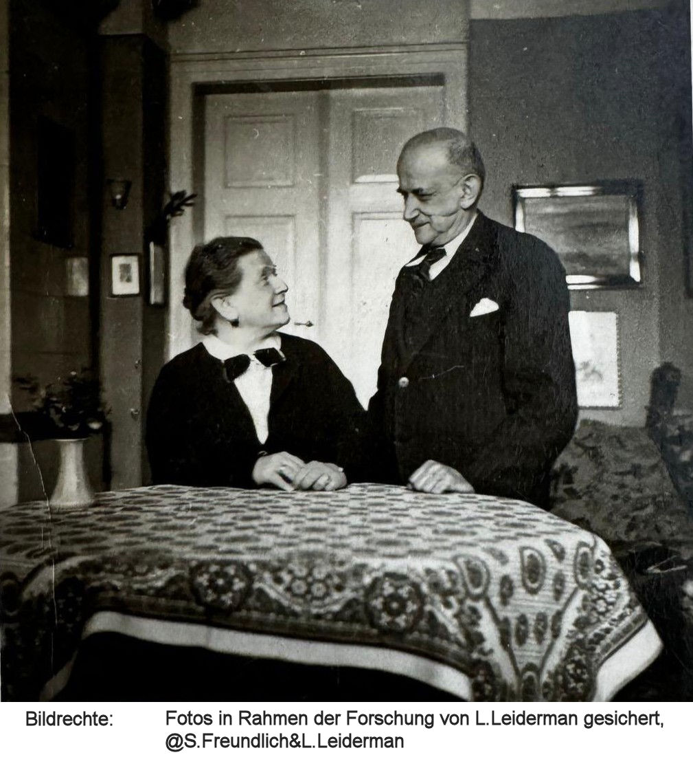 Schwarzweißbild - Henriette sitz an einem Tisch in einem Wohnzimmer, Felix steht neben ihr und sie schauen sich an. Bildreche Fotos in Rahmen der Forschung von L.Leiderman gesichert, @S.Freundlich&L.Leiderman