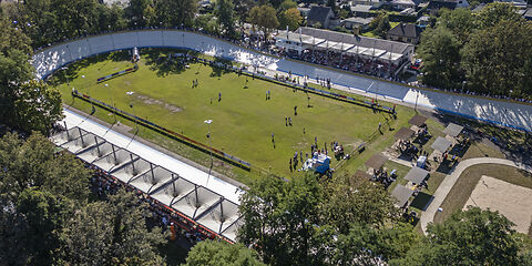 Blick von oben auf das neu sanierte Rad- und Reitstadion: Außen ist die Fahrbahn und in der Mitte befindet sich grüner Rasen, auf dem sich Menschen befinden. Im oberen und unteren Bereich des Bildes befinden sich die Tribühnen.
