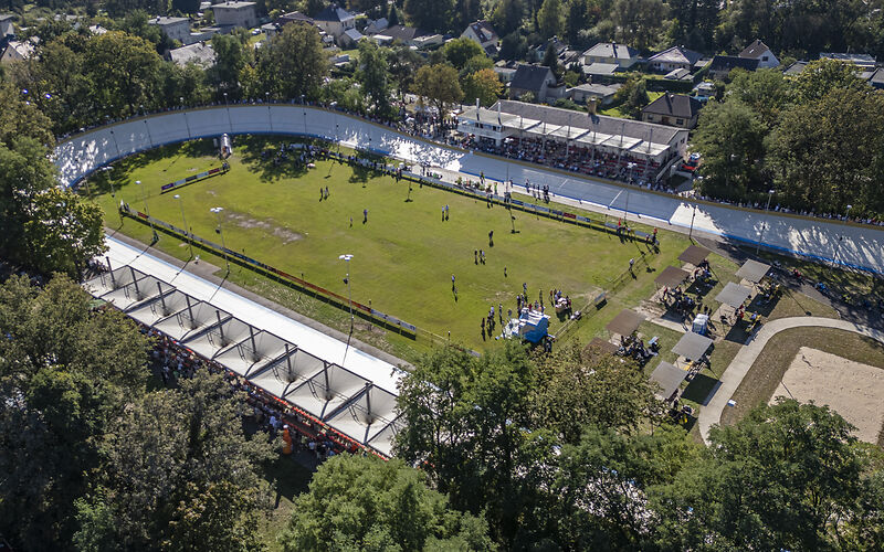 Blick von oben auf das neu sanierte Rad- und Reitstadion: Außen ist die Fahrbahn und in der Mitte befindet sich grüner Rasen, auf dem sich Menschen befinden. Im oberen und unteren Bereich des Bildes befinden sich die Tribühnen.