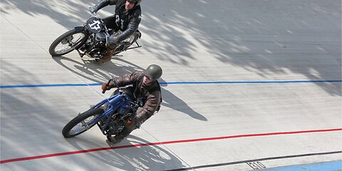 Auf der grauen Rennbahn fahren zwei Motoradfahrer in dunkler Kleidung nebeneinander.