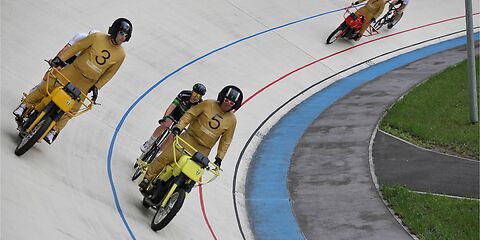 Auf der grauen Rennbahn fahren 3 Gespanne bestehend aus jeweils einem Schrittmacher (der vordere im Gespann) und einem Steher (hintere Teil des Gespanns). Die Schrittmacher tragen dunkelgelbe Anzüge mit Startnummern.