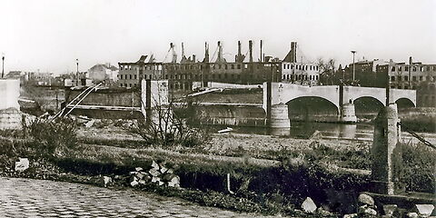 Schwarz-weiß Foto: Blick auf die zerstörte Brücke, im Hintergrund stehen mehrere Ruinen von Häusern, denen die Dächer und Fensterscheiben fehlen.