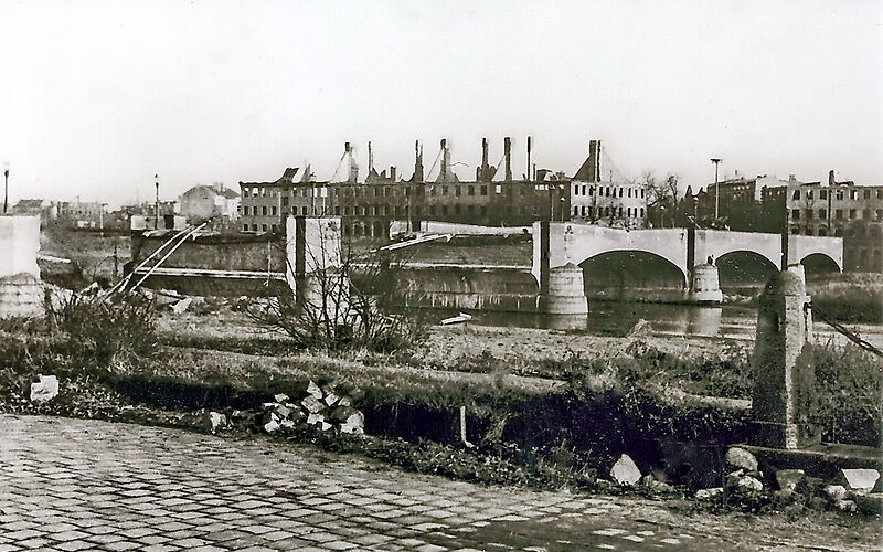 Schwarz-weiß Foto: Blick auf die zerstörte Brücke, im Hintergrund stehen mehrere Ruinen von Häusern, denen die Dächer und Fensterscheiben fehlen.