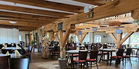 Das Foto zeigt den Gastraum des Restaurants im Gut Neu Sacro. Mehrere Tische sind schön eingedeckt und laden zum Verweilen ein. Sichtbare Holzbalken an der Decke und im Raum verleihen dem Restaurant eine warme, rustikale Atmosphäre.