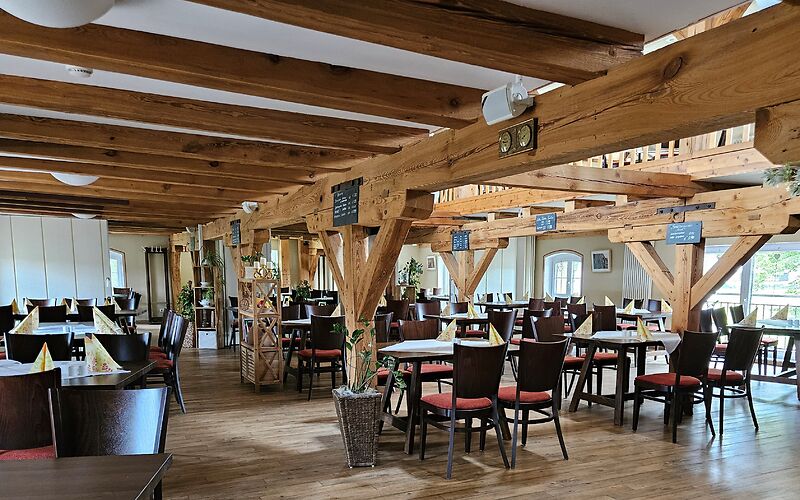Das Foto zeigt den Gastraum des Restaurants im Gut Neu Sacro. Mehrere Tische sind schön eingedeckt und laden zum Verweilen ein. Sichtbare Holzbalken an der Decke und im Raum verleihen dem Restaurant eine warme, rustikale Atmosphäre.
