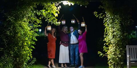 Eine Nachtaufnahme zeigt vier bunt gekleidete Personen mit leuchtenden Lampions unter einer beleuchteten Rosenpergola. Die stimmungsvolle Beleuchtung verleiht der Szene eine besondere Atmosphäre.