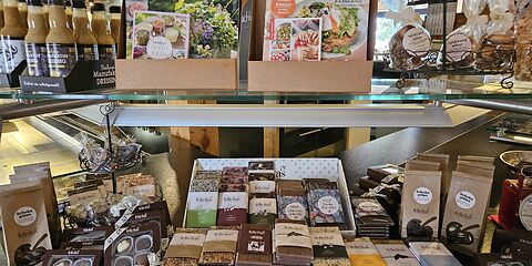 Das Foto zeigt den Hofladen im Gut Neu Sacro. Im Vordergrund befindet sich eine Verkaufsvitrine mit regionalen Produkten. Zu sehen sind unter anderem Schokolade in verschiedenen Geschmacksrichtungen und Pralinen der Confiserie Felicitas sowie Dressing und Heidesand-Gebäck.