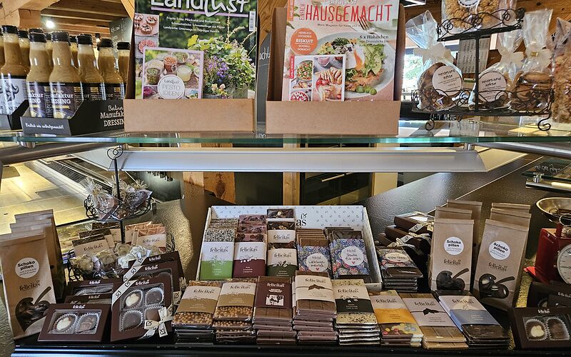 Das Foto zeigt den Hofladen im Gut Neu Sacro. Im Vordergrund befindet sich eine Verkaufsvitrine mit regionalen Produkten. Zu sehen sind unter anderem Schokolade in verschiedenen Geschmacksrichtungen und Pralinen der Confiserie Felicitas sowie Dressing und Heidesand-Gebäck.