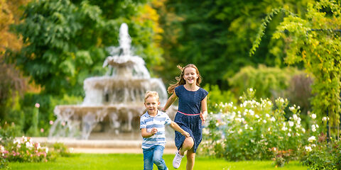 Das Foto zeigt ein Mädchen und einen Jungen, die lächelnd durch eine Wiese im Neurosengarten laufen. Im Hintergrund sind die Kaskaden mit fließendem Wasser sowie grün und orange gefärbte Bäume zu sehen.