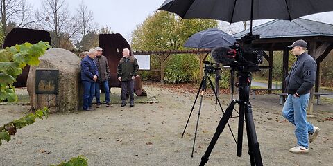 Die Vorbreitungen für den anstehenden Dreh des Interviews laufen. Die Kameras stehen schon bereit. Diese sind mit Schirmen abgedeckt, weil es regnet. Die 3 Interviewten stehen neben dem Denkstein von Weißagk und werden vom Technicker gerade verkabelt.