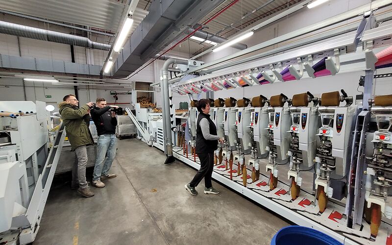 Wir befinden und in der Spinnerei in Forst. In der Maschienehalle steht eine Mitarbeiterin vor einer Maschine, sie erklärt wie diese funktioniert. Dabei wird sie von 2 Männern gefilmmt. Beide halten je eine schwarze Kamera in der Hand.