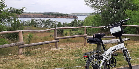 Ein weißen Fahrrad steht auf reinem Radweg vor einem Holzzaun. Im Hintergrund ist eine Wiese und der Klinger See zu sehen.