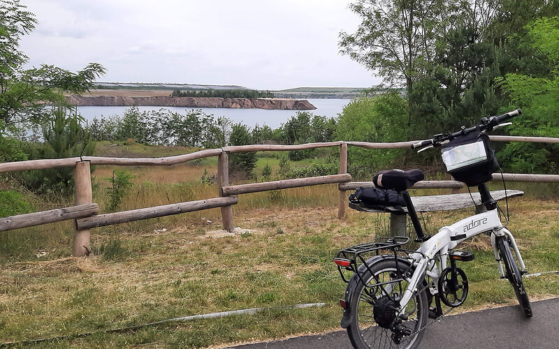 Ein weißen Fahrrad steht auf reinem Radweg vor einem Holzzaun. Im Hintergrund ist eine Wiese und der Klinger See zu sehen.