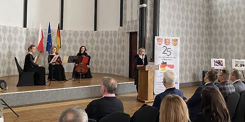 Die Bürgermeisterin Simone Taubenek steht am Rednerpult rechts neben der Bühne neben einem Banner auf dem die Wappen der 3 Städte, eine große 25 und weitere Informationen auf polnisch stehen. Auf der Bühne sitzen 3 Musikerinnen mit Streichinstrumenten.