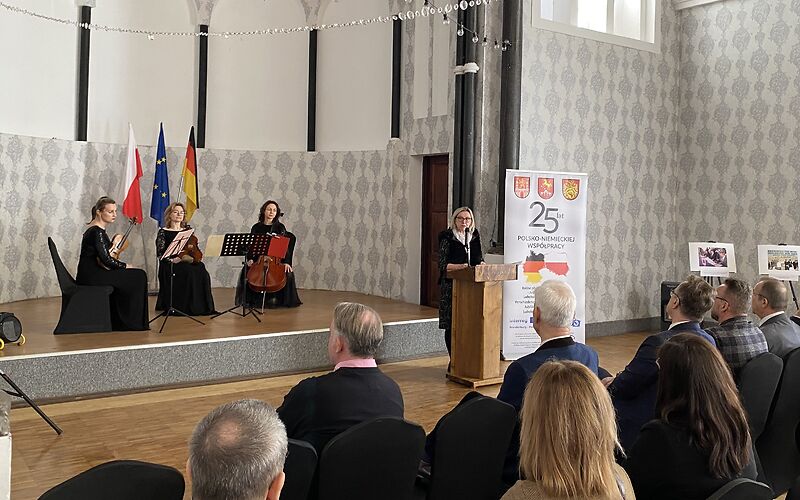 Die Bürgermeisterin Simone Taubenek steht am Rednerpult rechts neben der Bühne neben einem Banner auf dem die Wappen der 3 Städte, eine große 25 und weitere Informationen auf polnisch stehen. Auf der Bühne sitzen 3 Musikerinnen mit Streichinstrumenten.