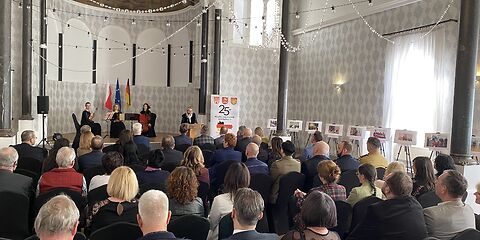 Blick von hinten auf die Gäste der Festveranstaltung: Im Saal sind Stuhlreihen gestellt, auf denen die Gäste sitzen. Rechts sind Bilder aufgestellt. Oben im Bild ist ein goldener Kronleuchter, von dem mehrere Perlenketten zur Seite weg führen, zu sehen.