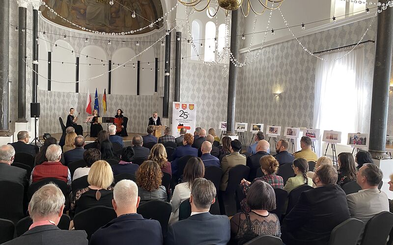 Blick von hinten auf die Gäste der Festveranstaltung: Im Saal sind Stuhlreihen gestellt, auf denen die Gäste sitzen. Rechts sind Bilder aufgestellt. Oben im Bild ist ein goldener Kronleuchter, von dem mehrere Perlenketten zur Seite weg führen, zu sehen.