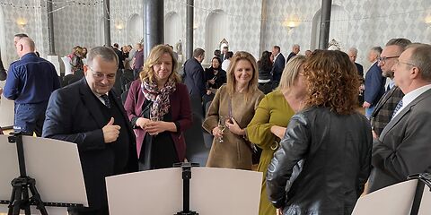 Die Besucher der Festveranstaltung schauen die ausgestellten Bilder an. Die Bilder sind nur von hinten zu sehen (weiße Rückseite). Links im Bild steht der Verwaltungsvorstand Herr Zuber. Im Hintergrund sind weitere Veranstaltungsgäste zu sehen, die miteinander reden.