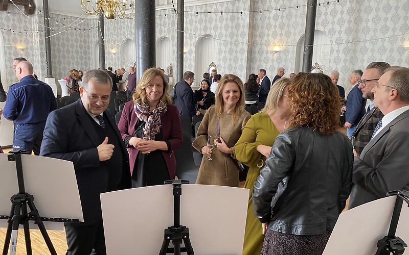 Die Besucher der Festveranstaltung schauen die ausgestellten Bilder an. Die Bilder sind nur von hinten zu sehen (weiße Rückseite). Links im Bild steht der Verwaltungsvorstand Herr Zuber. Im Hintergrund sind weitere Veranstaltungsgäste zu sehen, die miteinander reden.