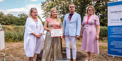 Rosenkönigin Stefanie III., Beata Kowalczuk, Janusz DudojÄ‡ und Simone Taubenek stehen vor einem der neu geplanzten Bäume. Stefanie die III. hält eine Infotafel zur Baumpflanzung in den Händen. Rechts steht ein Banner zum Fördermittelprojekt Interreg.
