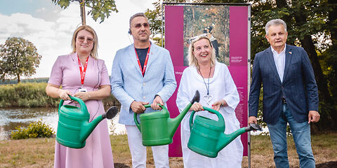 Beata Kowalczuk, Janusz DudojÄ‡ und Simone Taubenek stehen mit jeweils einer grünen Gieskanne in der Hand vor den neu gepfanzten Bäumen.
