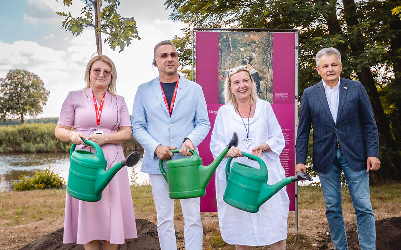 Beata Kowalczuk, Janusz DudojÄ‡ und Simone Taubenek stehen mit jeweils einer grünen Gieskanne in der Hand vor den neu gepfanzten Bäumen.