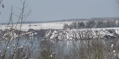 schneebedeckte Böschungen hinter dem dem Klinger See, im Vordergrund blätterlose Bäume