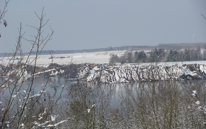 schneebedeckte Böschungen hinter dem dem Klinger See, im Vordergrund blätterlose Bäume