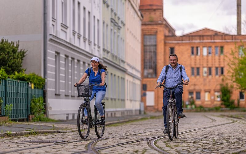 Ein Mann und eine Frau fahren auf einer gepflasterten Straße nebeneinander Fahrrad. Auf der Straße sind Schienen verlegt. Links steht ein Wohnhaus und im Hintergrund ist eine altes Fabrikgebäude aus Backstein zu sehen.