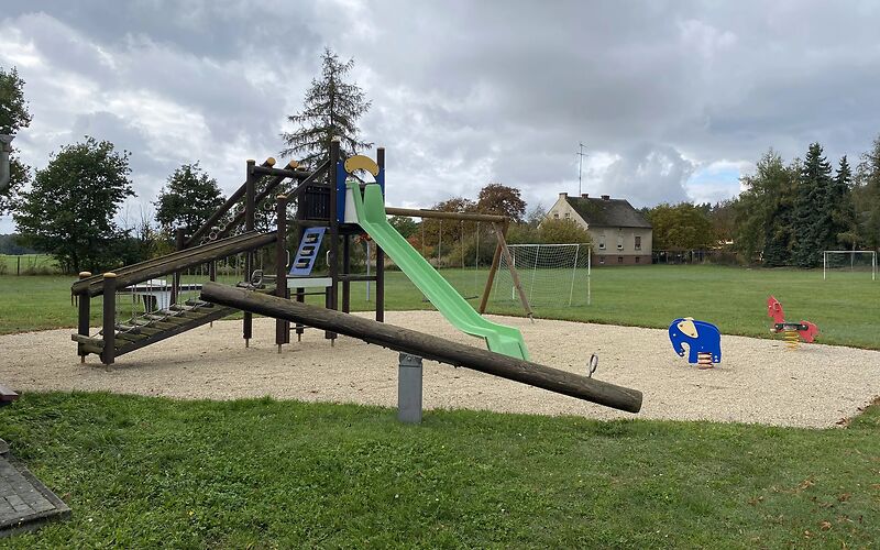 Auf einer Sandfläche stehen ein Klettergerüst aus Holz, an dem eine grüne Rutsche befestigt ist und zwei Federwippen (rotes Pferd und blauer Elefant). Davor steht auf der Wiesen eine Holzwippe.