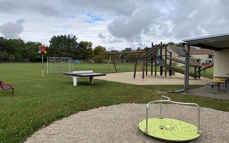 Vorn ist ein Karussel (Drehscheibe mit drei zur Mitte gebogenen Metallstangen) eine Tischtennisplatte, ein Klettergerüst und ein Fußballtor.
