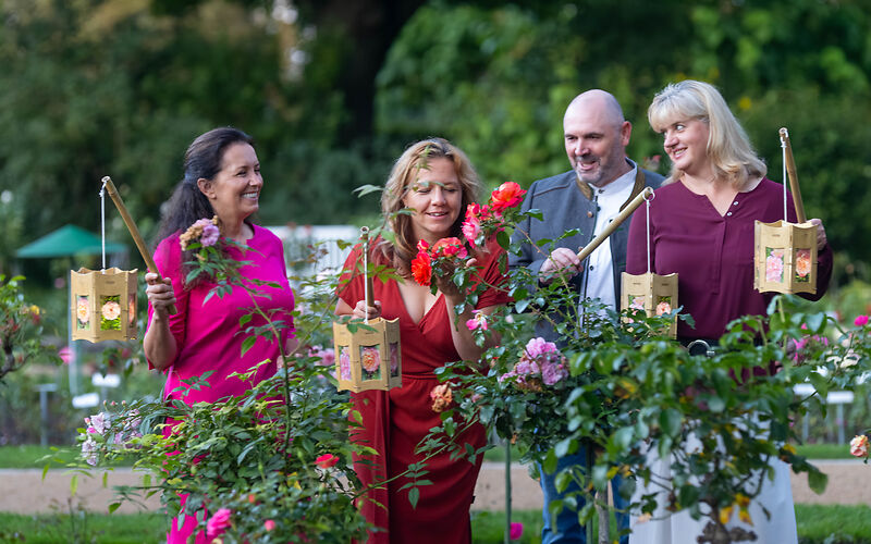Drei Frauen und ein Mann erkunden mit leuchtenden Lampions den Hochstamm- und Edelrosengarten während der romantischen Nachtführung.