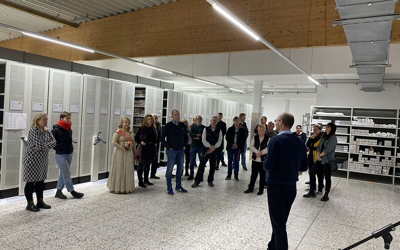 Blick in das Magazin: Schiebbare Regale mit Archivgut und offene Lagerregale. Im Raum steht die Gruppe Besucher.