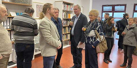 Im Lesesaal stehen die Besucher der Archiveröffnung. An der Wand links befinden sich Regale mit Büchern/Ordnern.