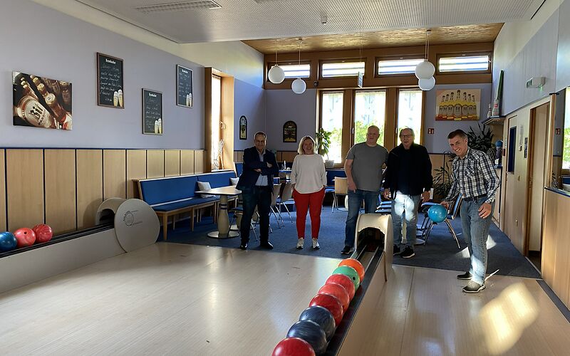 Die Gruppe steht in einem hellen Bowlingraum, eine Person wirft eine blaue Kugel auf der Bahn