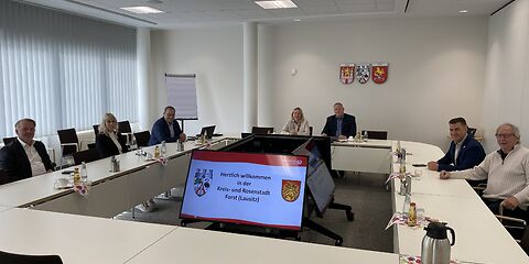 Die Verwaltungsvorstände und die Bürgermeisterin der Stadt Forst (Lausitzen) sitzen mit den Gästen aus Wermelskirchen im Sitzungssaal der Stadt an einer rechteckigen Tafel aus weißen Tischen. Im Hintergrund hängen die Wappen der 3 Partnerstädte an der Wand.