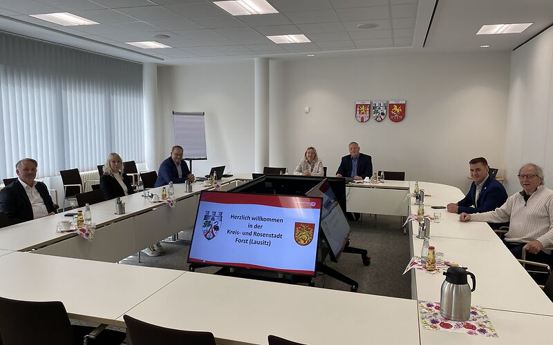 Die Verwaltungsvorstände und die Bürgermeisterin der Stadt Forst (Lausitzen) sitzen mit den Gästen aus Wermelskirchen im Sitzungssaal der Stadt an einer rechteckigen Tafel aus weißen Tischen. Im Hintergrund hängen die Wappen der 3 Partnerstädte an der Wand.