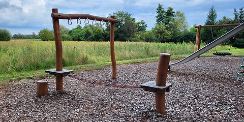 Spielgerät aus Holz mit drei verbundenen Seilen, die zwischen zwei Plattformen gespannt sind sowie einem waagerecht auf zwei Säulen liegenden Holzbalken, an dem mehrere Griffe befestigt sind.