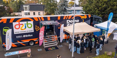 Stand auf dem Tag des offenen Unternehmens: Ein LKW, der durch eine Treppe mittig begehbar ist mit der Aufschrift "deine Zukunft ist bunt", davor stehen 2 Pavillione. Unter dem vorderen Pavillion steht eine große Gruppe Jugendlicher.