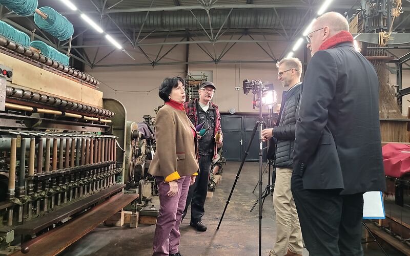 Im Museumsdepot filmen 2 Peronen ein Interview mit einer Dame. Diese steht vor einer Maschiene mit verschiedenen Spulen. Des Weiteren steht ein Mitarbeiter vom Museum mitdabei und hält 3 Spulen in den Farben galb, lila und blau in der Hand.