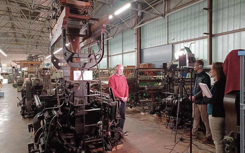 Im Museums Depot neben den alten Maschinen des Musems wird ein älterer Mann gefilmt und interviewt. Er trägt einen roten Poluver und eins schwarze Hose. Hinter der Kamera stehen 2 Personen, diese sind für die Regie zuständig.