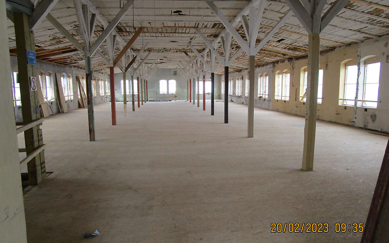 Zu sehen ist der fertiggestellte Fußboden. Auf der linken und rechten Seite, sowie in der Mitte befinden sich Industriefenster. Eine Reihe von Stützpfeilern ist auf der linken und rechten Seite zu sehen.