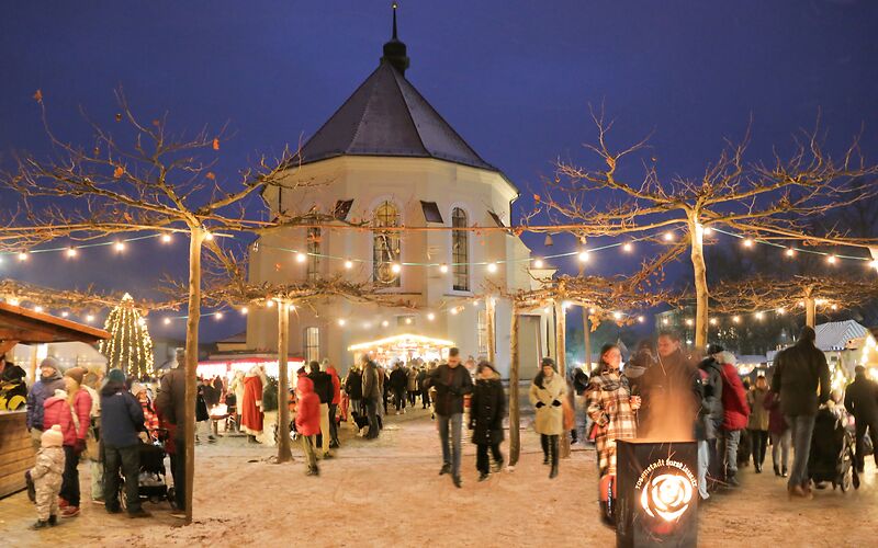 auf dem Forster Weihnachtsmarkt, mit Schneegestöber sind im Vordergrund unter dem mit Licherketten beleuchteten Platanenhain Menschen zu sehen, die an einer unter anderem an einer Feuerschale stehen. Im Hintergrund ist das Kirchenschiff der St. Nikolai zu sehen.