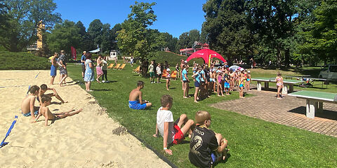 Die Kinder und Jugendlichen stehen bei den Tischtennissplatten. Einige sitzen auf dem Beachvolleyballfeld oder auf dem Gras.