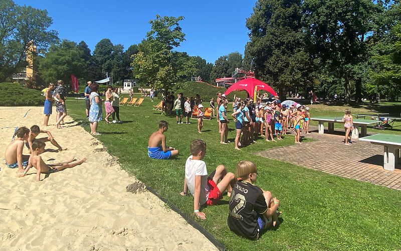 Die Kinder und Jugendlichen stehen bei den Tischtennissplatten. Einige sitzen auf dem Beachvolleyballfeld oder auf dem Gras.