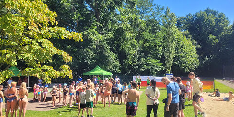 Viele Kinder und Jugendliche stehen mit dem Rücken zur Kamera auf einer Wiese. Vor ihnen steht ein Moderator mit einem Mikrofon, um etwas aanzukündigen.