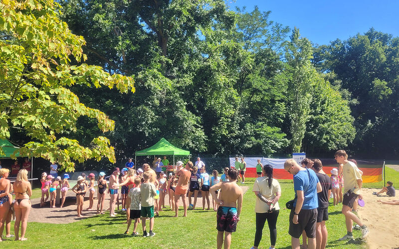 Viele Kinder und Jugendliche stehen mit dem Rücken zur Kamera auf einer Wiese. Vor ihnen steht ein Moderator mit einem Mikrofon, um etwas aanzukündigen.