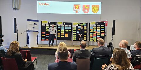 Auf der Bühne stehen ein Man und eine Frau, hinter ihnen sind 4 Pinnwände, an denen bunte beschriebene Zettel angepinnt sind. Vor der Bühne sitzen die Teilnehmenden in Stuhlreihen.