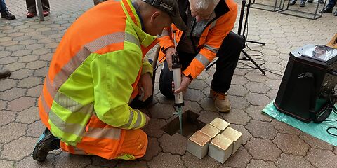 Zwei Männer bei der Einsetzung der Stolpersteine in den Gehweg. Die Steine liegen vor dem für sie ausgehobenen Loch. Einer der Männer hält eine Spritze mit Beton in der Hand.