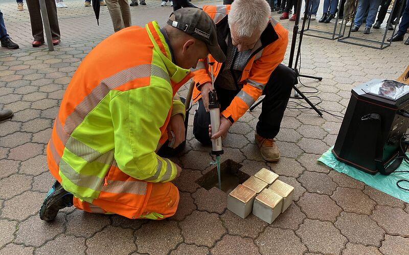 Zwei Männer bei der Einsetzung der Stolpersteine in den Gehweg. Die Steine liegen vor dem für sie ausgehobenen Loch. Einer der Männer hält eine Spritze mit Beton in der Hand.